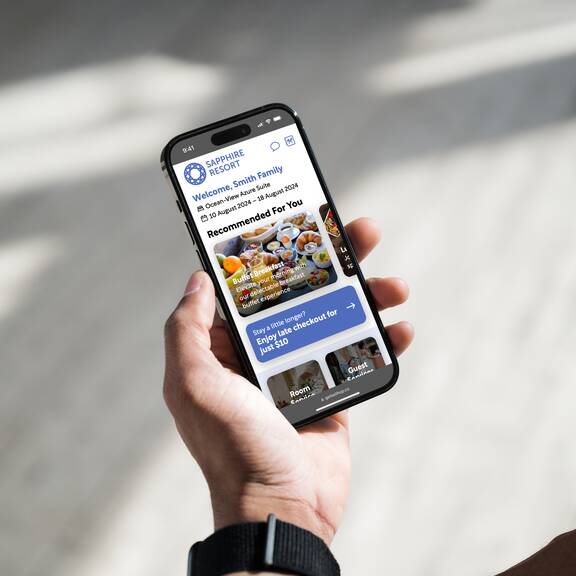 A guest holding a smartphone displaying the home screen of the Bellhop guest compendium system. Several recommendations are displayed at the top with a prominent contextual promotion for late checkout showing below, followed by a menu containing “Room Service”, “Guest Services”, and other items.